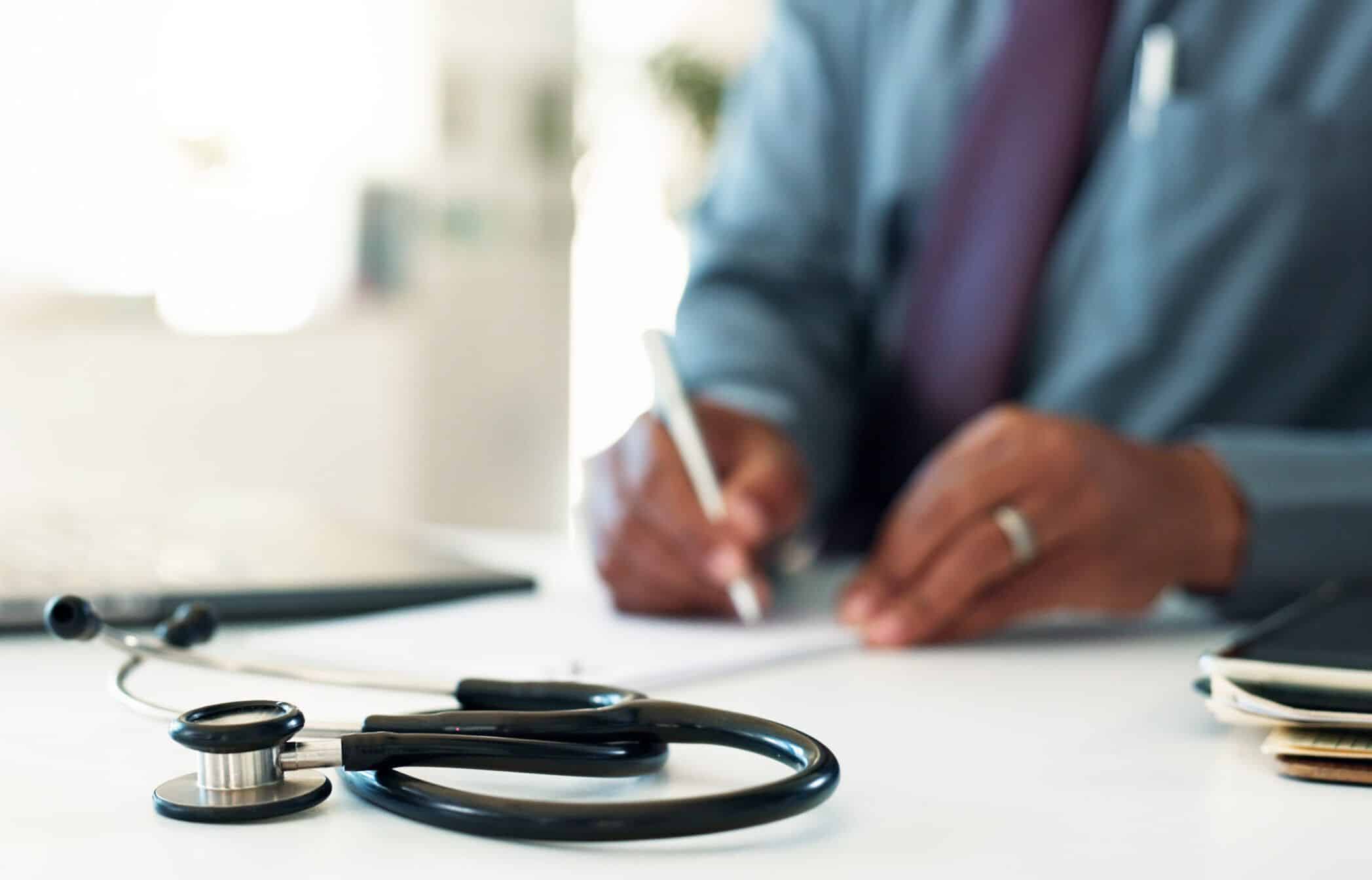 Manos de un doctor escribiendo documentación médica junto a un portátil